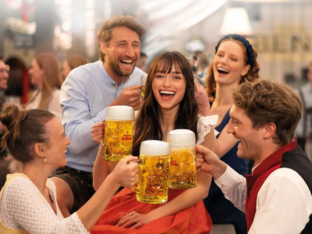 Fünf Personen stoßen lachend mit großen Bierkrügen an. Im Hintergrund sitzen weitere Gäste in einem Festzelt.