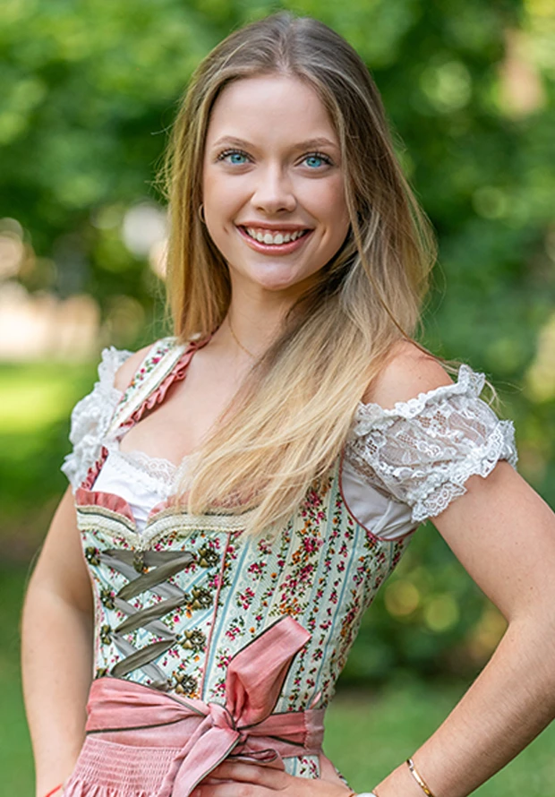 Eine lächelnde junge Frau trägt ein traditionelles Dirndl in einem Park.