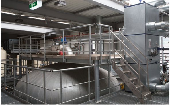 Ein Industrieraum mit großen Metalltanks und einer Stahlplattform, umgeben von Geländern und Rohren.