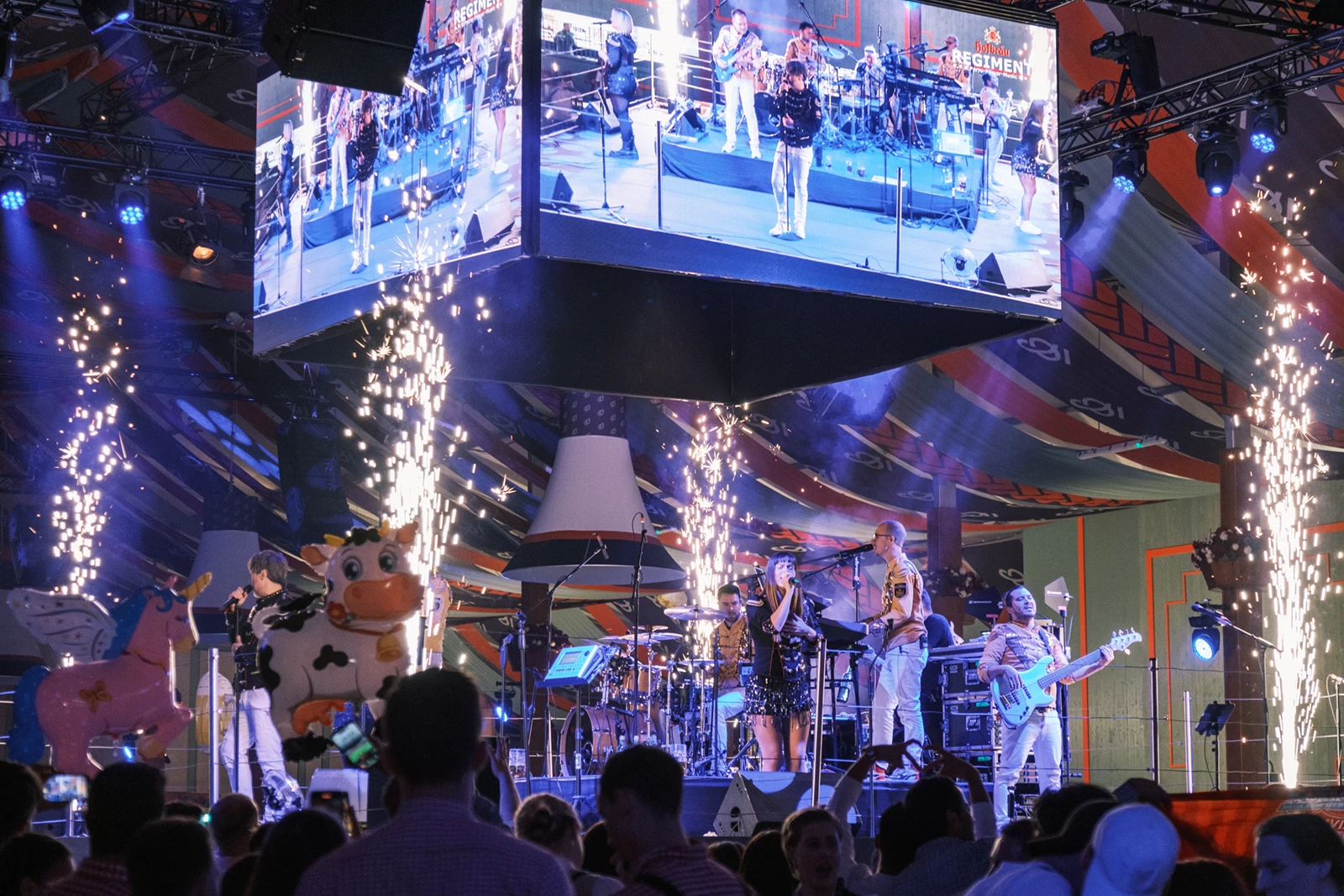 Eine Band spielt auf einer hell erleuchteten Bühne mit Pyrotechnik und großem Bildschirm. Im Hintergrund sind bunte Zeltdecken.