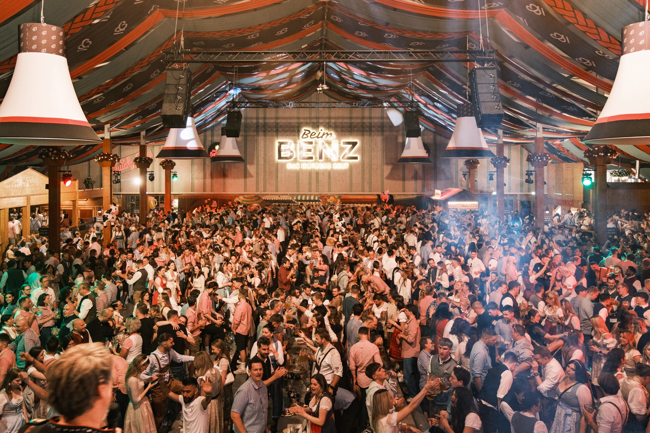 Viele Menschen in traditioneller Kleidung feiern dicht gedrängt im Benz Hofbräu Festzelt. Im Hintergrund ein leuchtendes Schild.