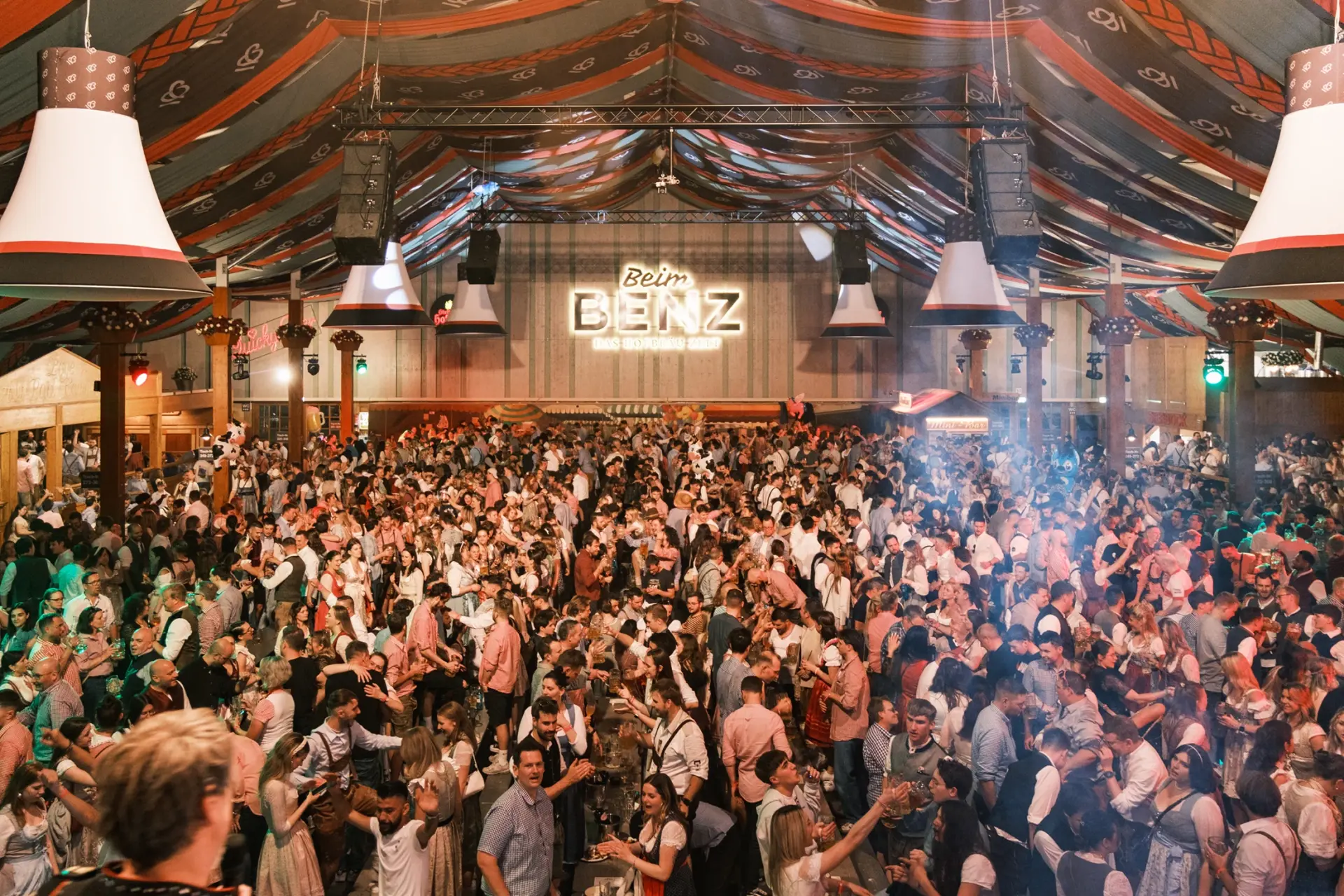 Viele Menschen in traditioneller Kleidung feiern dicht gedrängt im Benz Hofbräu Festzelt. Im Hintergrund ein leuchtendes Schild.