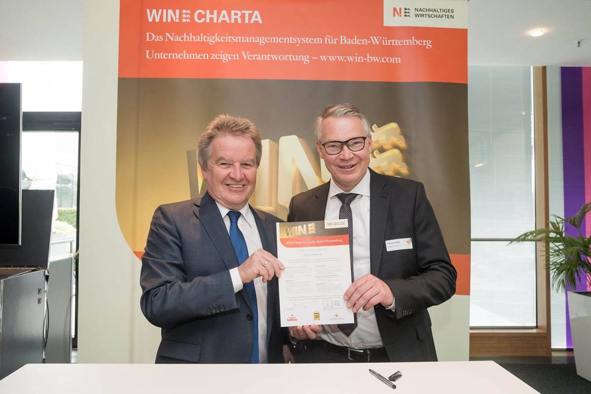 Zwei Herren in Anzügen halten stehend eine Urkunde in einem Büro.