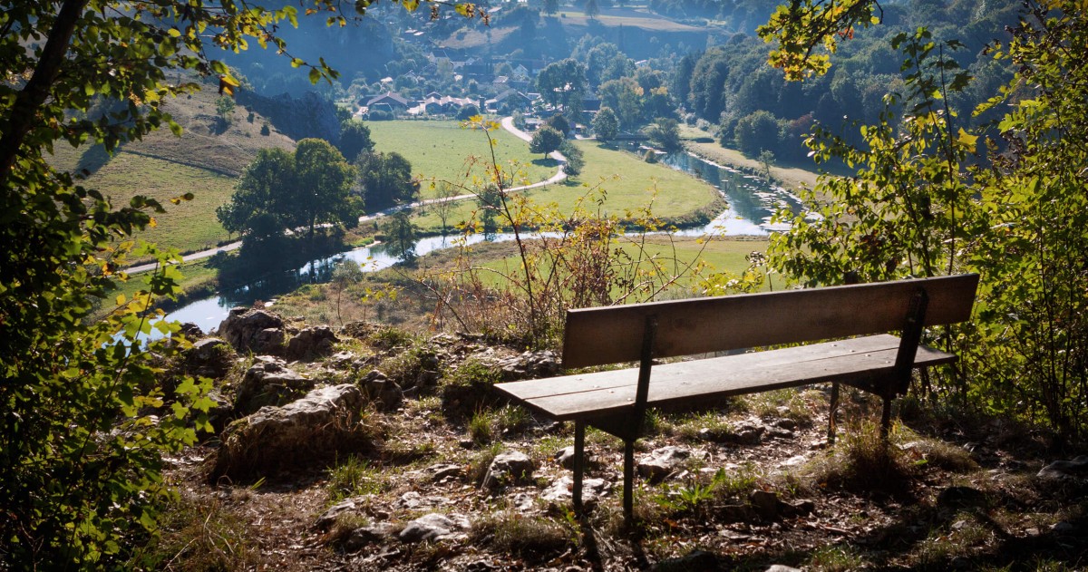 Eine Holzbank überblickt einen Fluss, der sich durch eine ländliche Landschaft schlängelt.