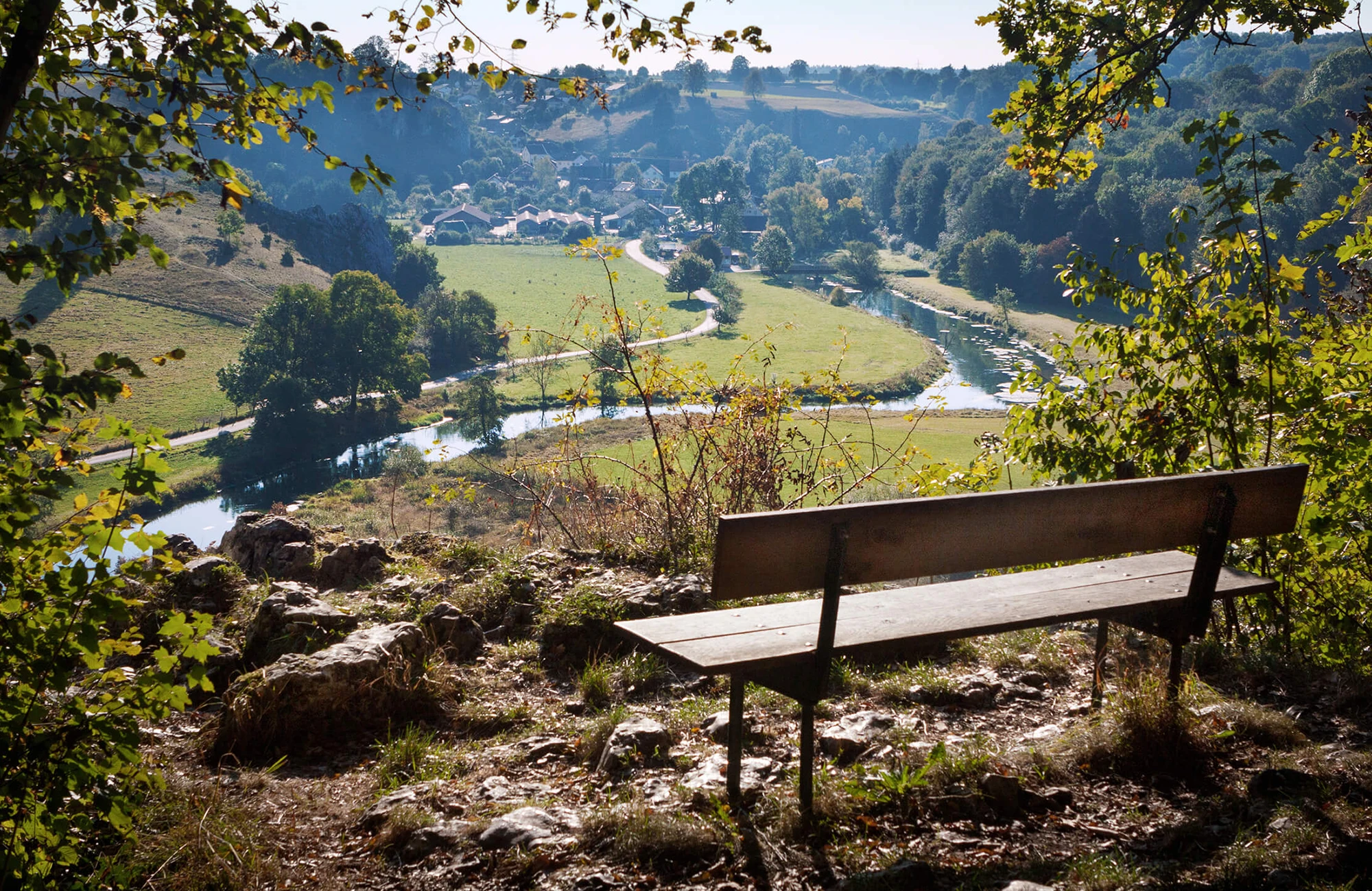 Holzbank auf erhöhtem Aussichtspunkt. Im Hintergrund ein Tal mit Fluss, Wiesen, Bäumen und verstreuten Häusern.