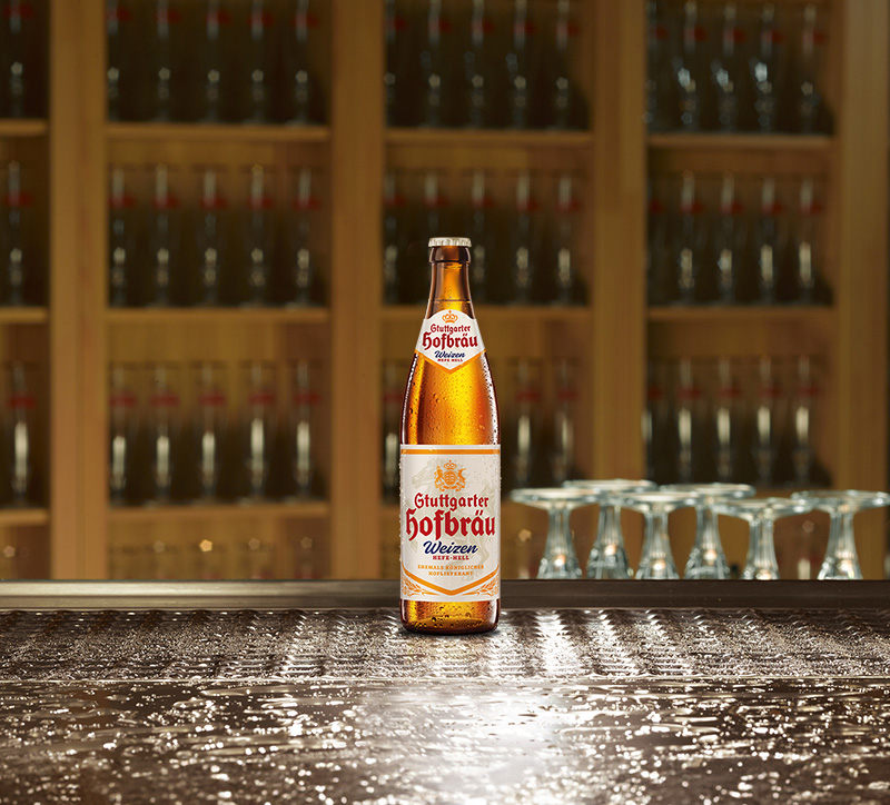 Eine Bierflasche steht auf einem nassen Holztisch, im Hintergrund sind Regale mit weiteren Flaschen.