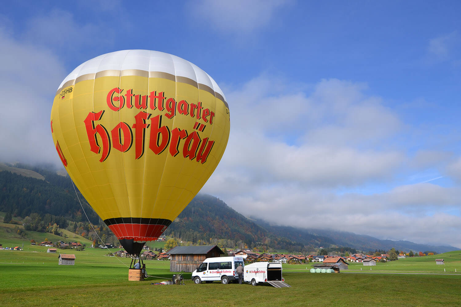 Ein großer Heißluftballon steht auf einer Wiese vor einer Bergkulisse, neben einem weißen Van.