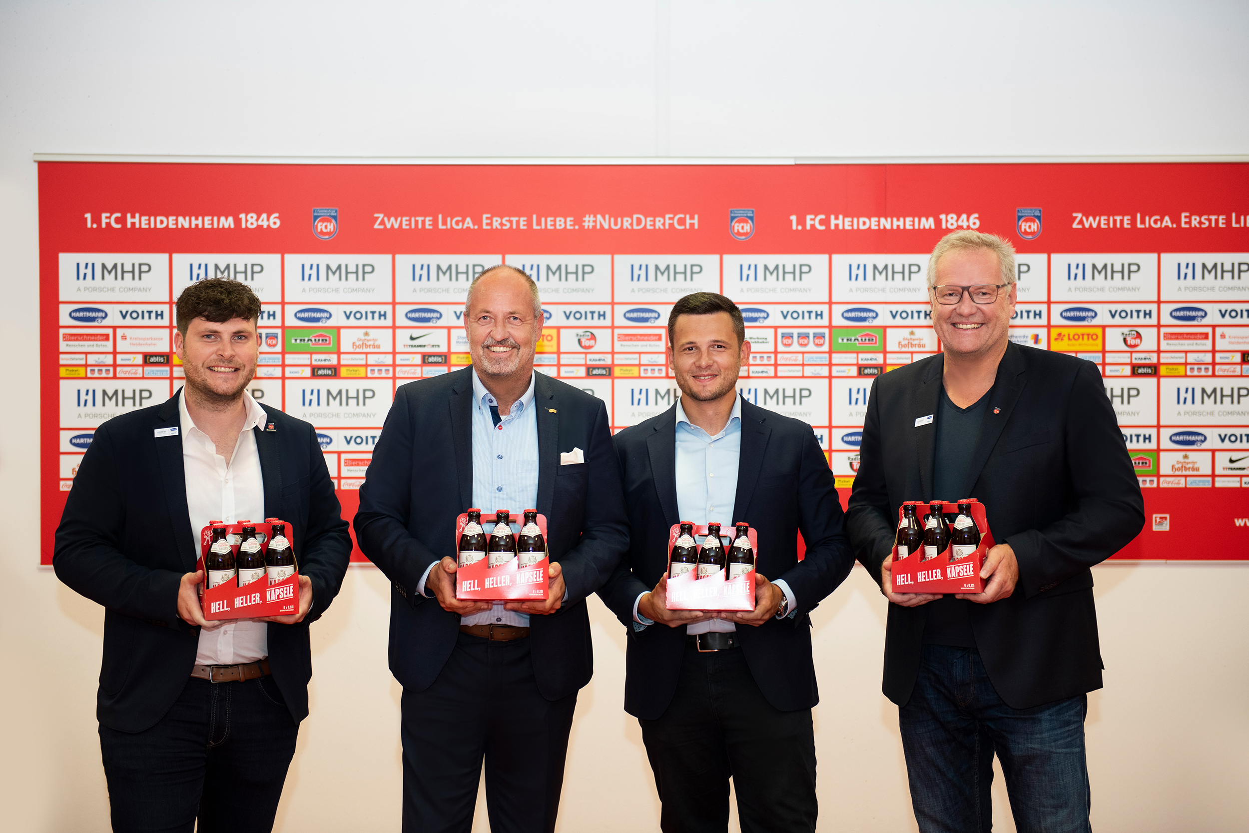Vier Männer stehen lächelnd nebeneinander vor einer Wand mit Logos und halten Bierpackungen.