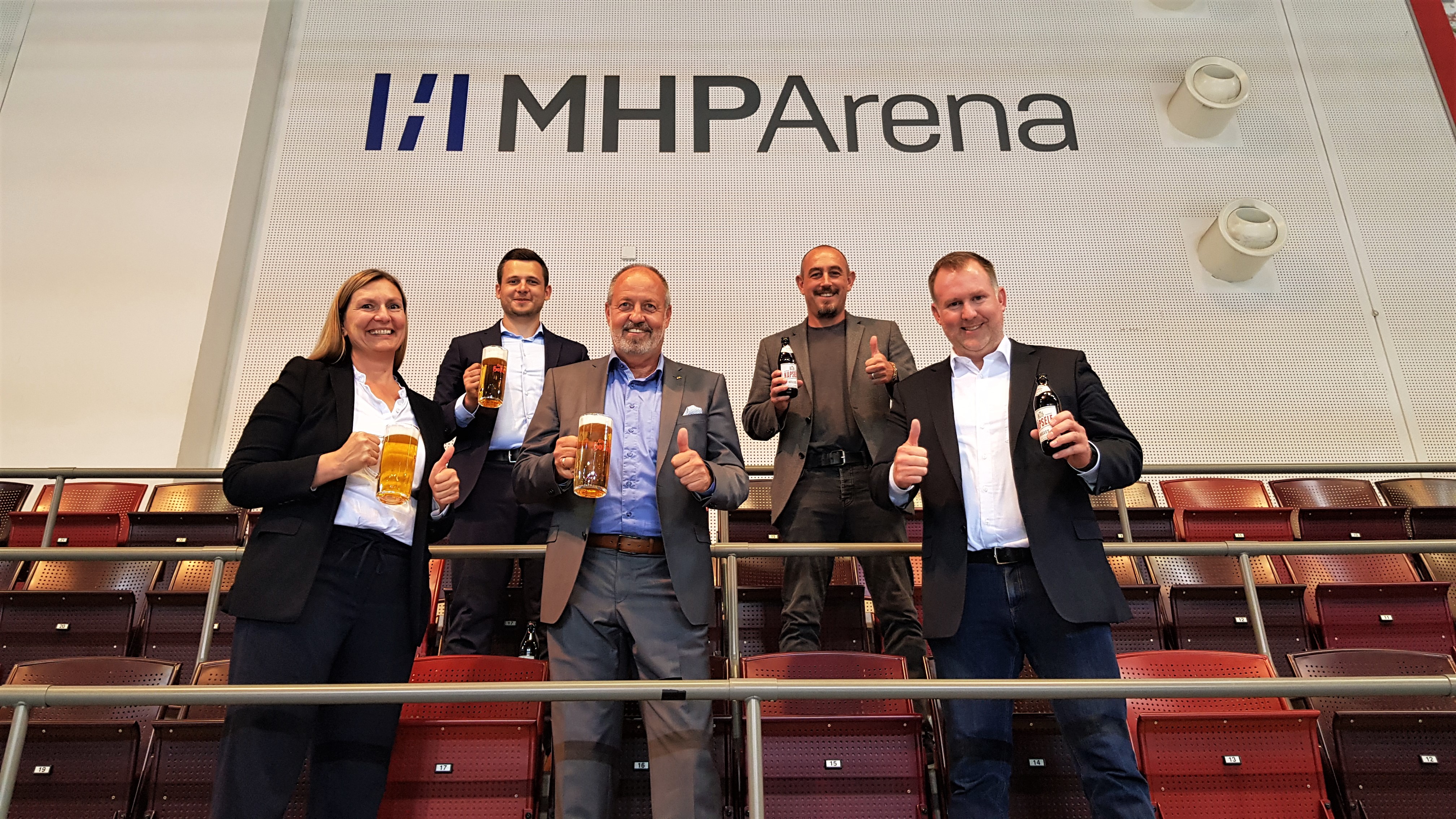 Fünf Personen heben in einer Arena lachend ihre Biergläser in einer Reihe.