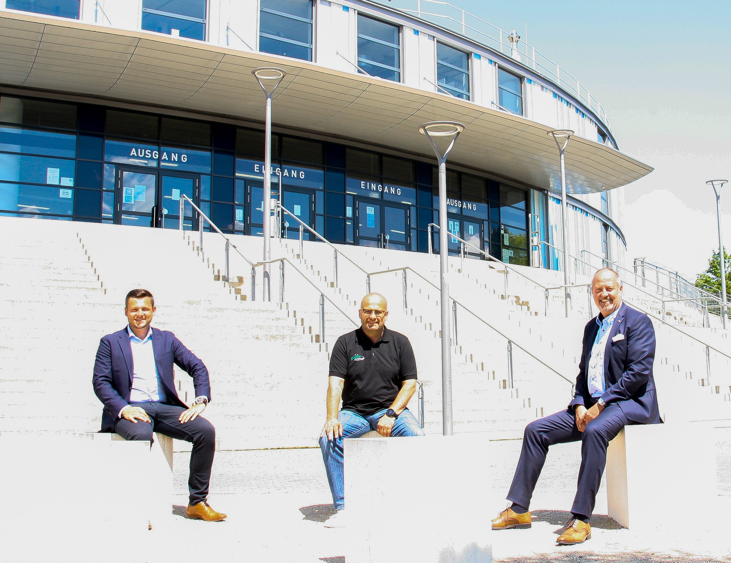 Drei Männer sitzen vor einem modernen Gebäude mit großen Fenstern und Türen beschriftet als Eingang und Ausgang.