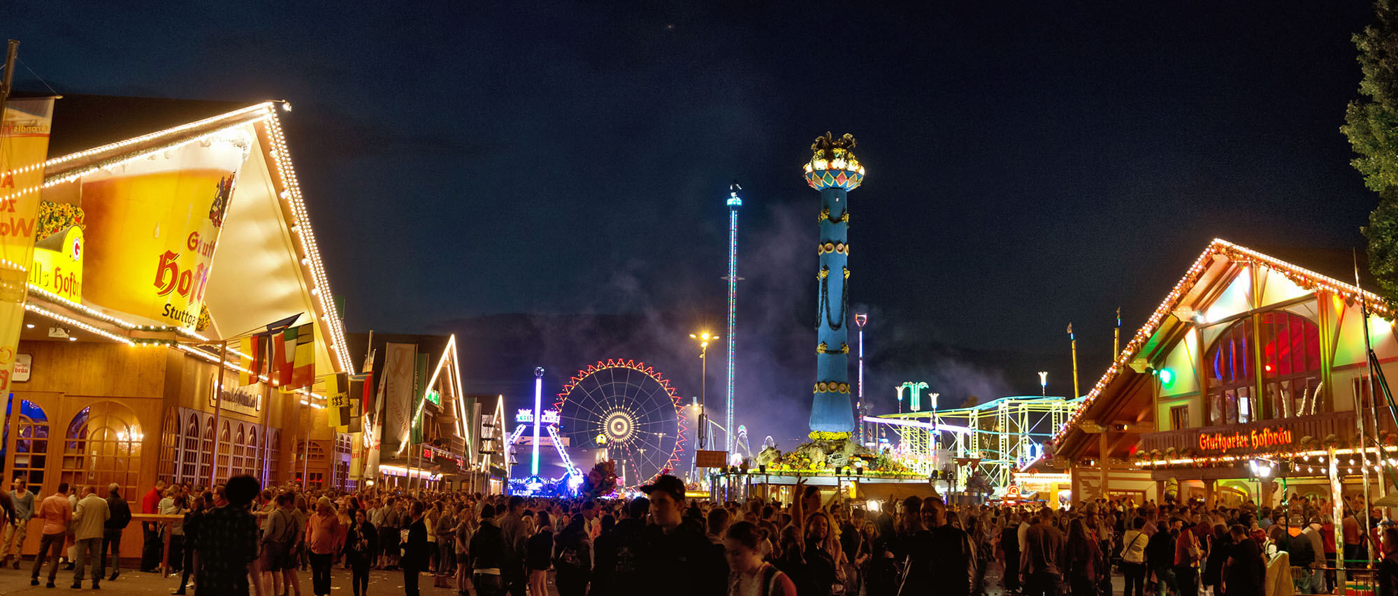 Eine nächtliche Kirmes mit beleuchteten Fahrgeschäften und Buden, Menschenmengen flanieren entlang der Stände.