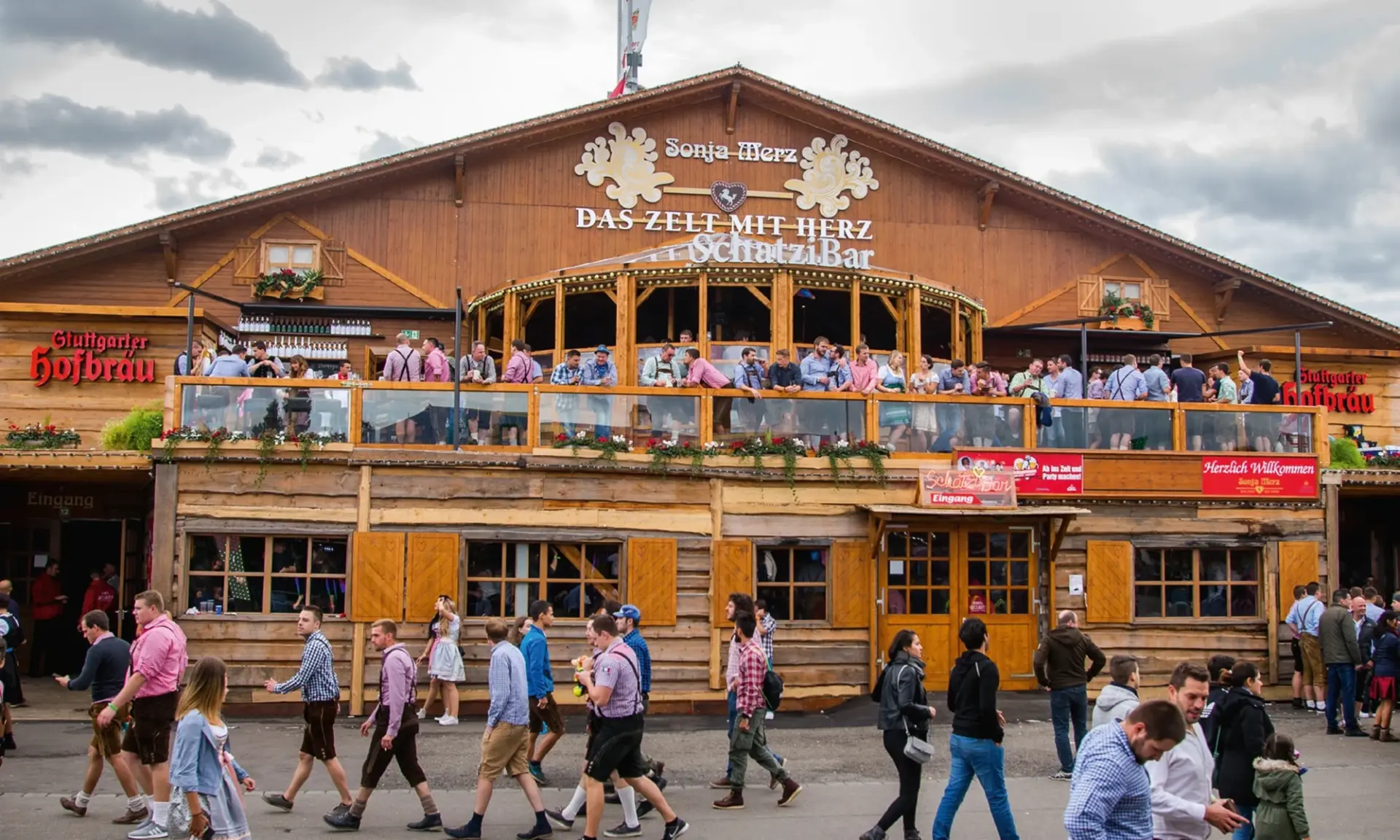 Menschen in traditioneller Kleidung gehen an einem großen, holzverkleideten Festzelt vorbei. Auf den Balkonen des Zelts befinden sich weitere Besucher.