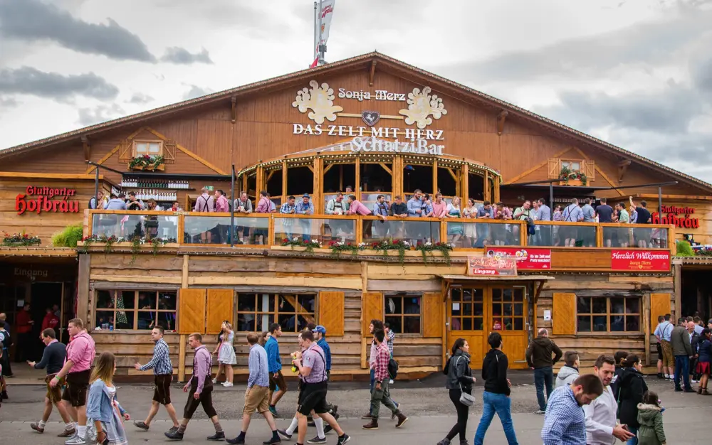 Menschen in traditioneller Kleidung gehen an einem großen, holzverkleideten Festzelt vorbei. Auf den Balkonen des Zelts befinden sich weitere Besucher.