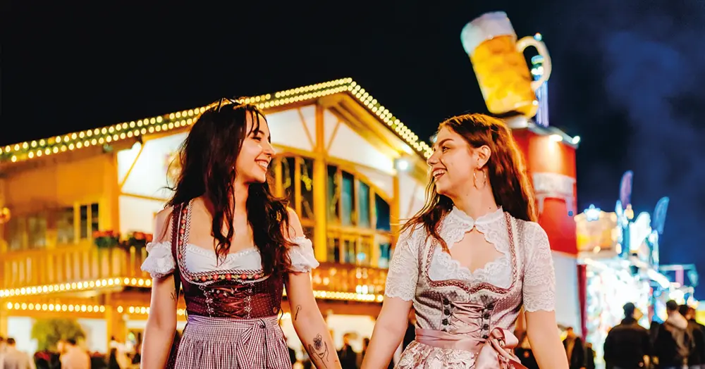 Zwei Frauen in Dirndlkleidern laufen nebeneinander und lächeln sich an. Im Hintergrund ein festlich beleuchtetes Gebäude.